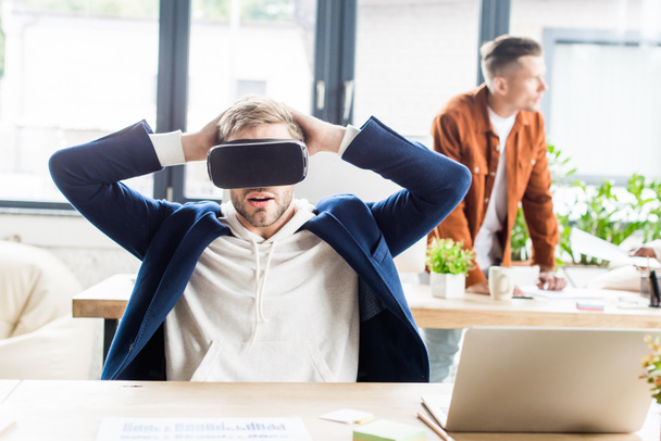 young businessman using vr headset and holding hands behind head while sitting at workplace in office - Photo, Image