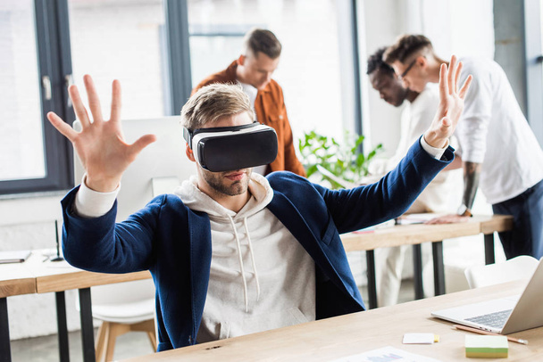 young businessman using vr headset and gesturing while multicultural colleagues working in office - Photo, Image