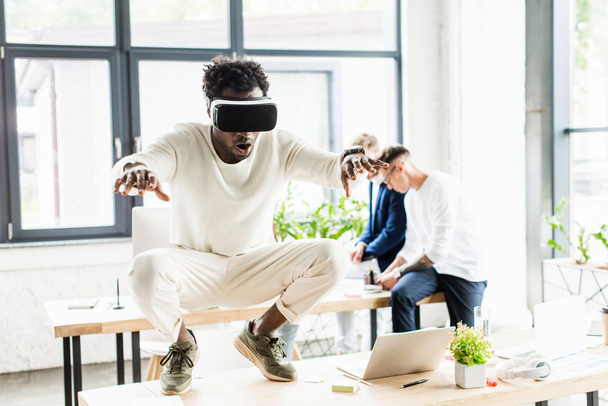 african american businessman using vr headset and squatting on desk while colleagues working in office - Foto, Imagem