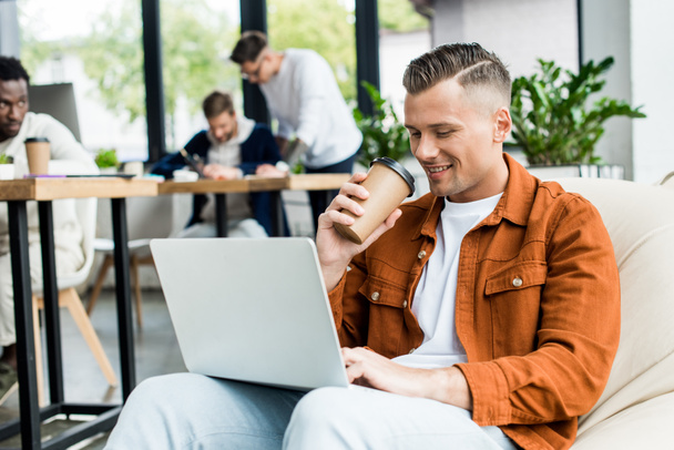 young businessman drinking coffee to go and using laptop while working in office near multicultural colleagues - Photo, Image