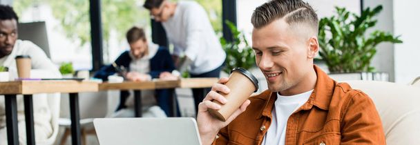 tiro panorâmico de jovem empresário beber café para ir e usar laptop enquanto trabalhava no escritório perto de colegas multiculturais
 - Foto, Imagem