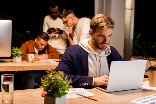 молодой, серьезный бизнесмен, работающий ночью на ноутбуке рядом с коллегами по мультикультурному бизнесу в офисе
 - Фото, изображение