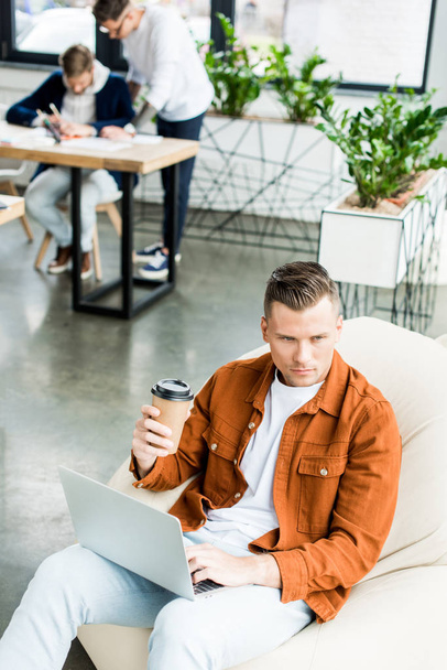 высокий угол обзора молодого бизнесмена с бумажной чашкой и с помощью ноутбука во время работы в офисе рядом с коллегами
 - Фото, изображение