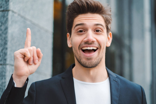 Portrait of young thinking pondering businessman having idea moment pointing finger up on office building background. Smiling happy student man showing eureka gesture. - Foto, Imagen