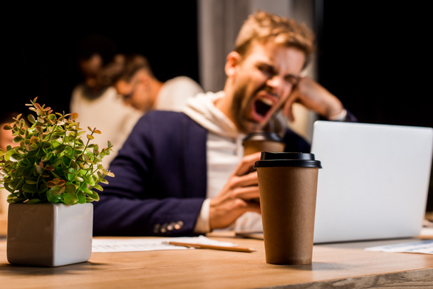 selective focus of disposable cup near exhausted businessman yawning while working at night in office - Photo, Image