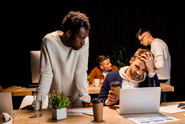 Afrikanisch-amerikanischer Geschäftsmann steht neben erschöpftem Kollegen und hält Kaffee to go, während er nachts im Büro arbeitet - Foto, Bild