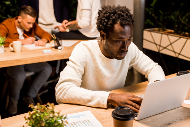 uomo d'affari afro-americano esausto che utilizza il computer portatile mentre lavora di notte in ufficio vicino ai colleghi
 - Foto, immagini