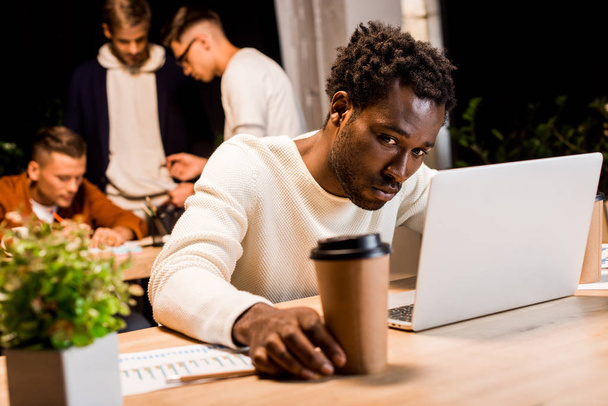 selective focus of exhausted african american businessman holding coffee to go while working at night in office near colleagues - Photo, Image