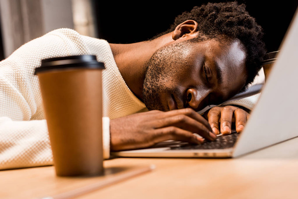 wybiórcze skupienie wyczerpanego afrykańskiego biznesmena śpiącego w miejscu pracy w pobliżu laptopa i jednorazowego kubka w nocy w biurze - Zdjęcie, obraz