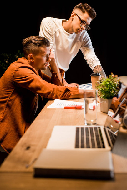 selektywne skupienie się młodego biznesmena stojącego w pobliżu wyczerpanego kolegi korzystającego w nocy z laptopa w biurze - Zdjęcie, obraz