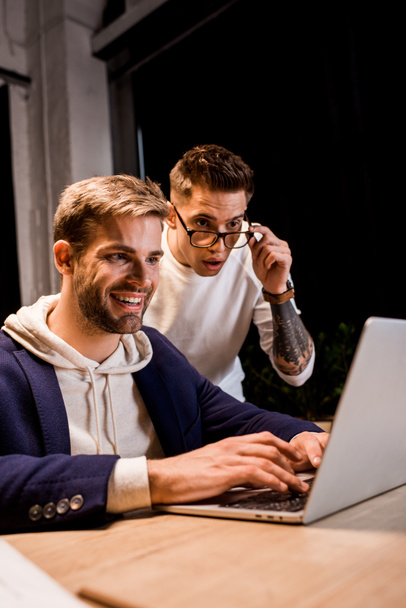 young businessman touching glasses while looking at laptop near happy colleague - Photo, Image