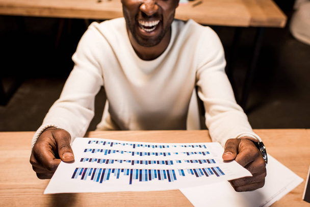 Afro-Amerikan iş adamının elinde grafikler ve çizelgeler olan bir kağıt tutarken çekilmiş görüntüsü.  - Fotoğraf, Görsel