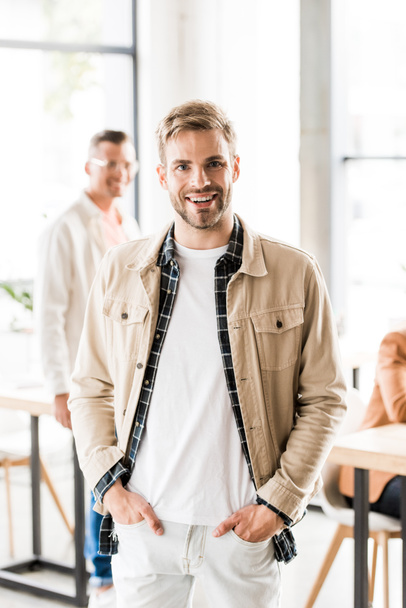 cheerful, young businessman standing with hands in pockets and smiling at camera  - Foto, afbeelding