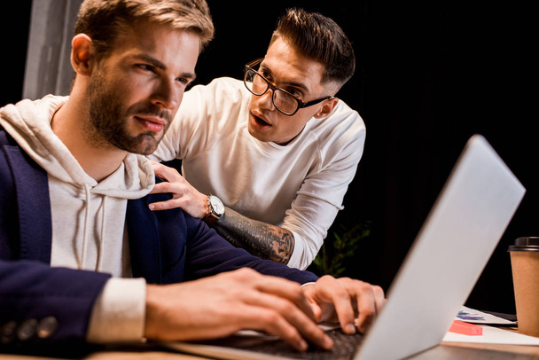 young businessman touching shoulder of colleague working on laptop at night in office - Фото, зображення