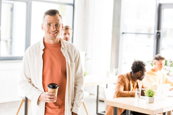 young, handsome businessman holding coffee to go and smiling at camera - Photo, Image