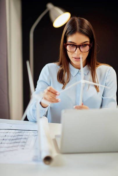 Charming hardworking brunette dressed in formal wear sitting at desk in office and holding windmill model. On desk is laptop. - Фото, зображення