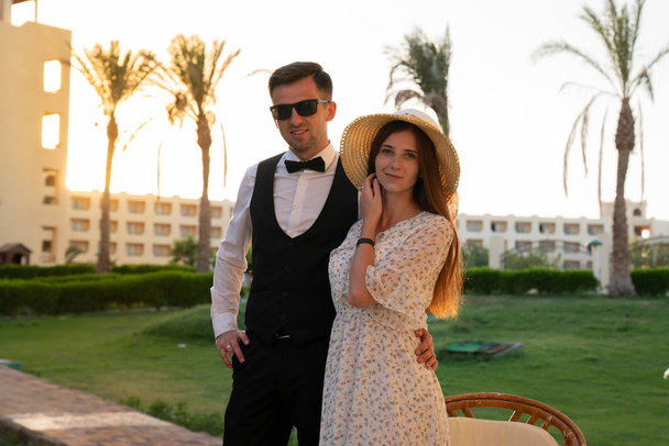 glückliches junges Paar, Mann in weißem Hemd und Sonnenbrille umarmt seine Freundin mit Hut und zartem Kleid, beide blicken in die Kamera, Sonnenuntergang im Hintergrund - Foto, Bild