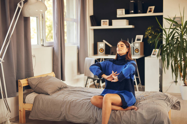 Beautiful Caucasian brunette in blue sweater sitting on bed in bedroom and stretching. Morning time. - Foto, Bild