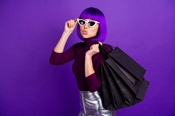 Красива стильна дама тримає багато пакетів в руках відправляє повітряний поцілунок, одягнений в модний одяг ізольований фіолетовий фон
 - Фото, зображення