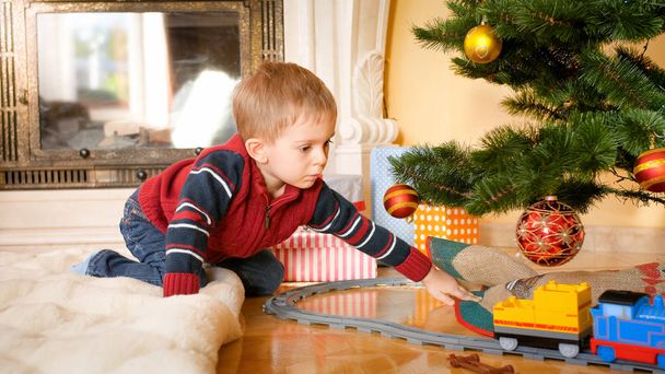 Portret van de kleine jongen zittend op de vloer in de woonkamer en kijken naar speelgoed trein op de spoorwegen onder kerstboom - Foto, afbeelding
