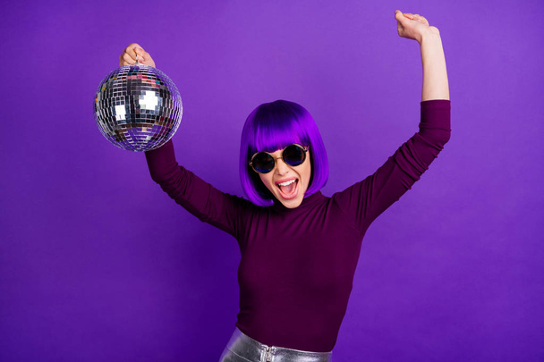 Portrait of cheerful girl with eyeglasses eyewear raising her arms screaming holding glowing ball wearing turtleneck isolated over purple violet background - Foto, immagini