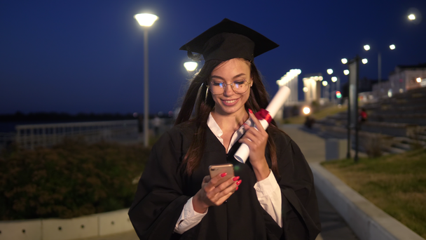Vysoká škola absolvent dívka chůze s diplomem s úsměvem a pomocí svého smartphonu. - Záběry, video