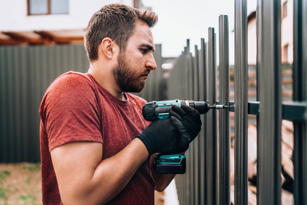 Construction worker using electrical screwdriver and mounting metal elements on fence - Foto, imagen