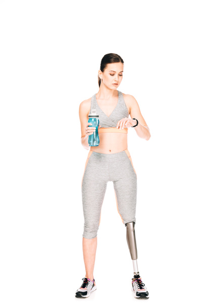 vue pleine longueur de la sportive avec prothèse jambe tenant bouteille de sport et en regardant smartwatch isolé sur blanc
 - Photo, image