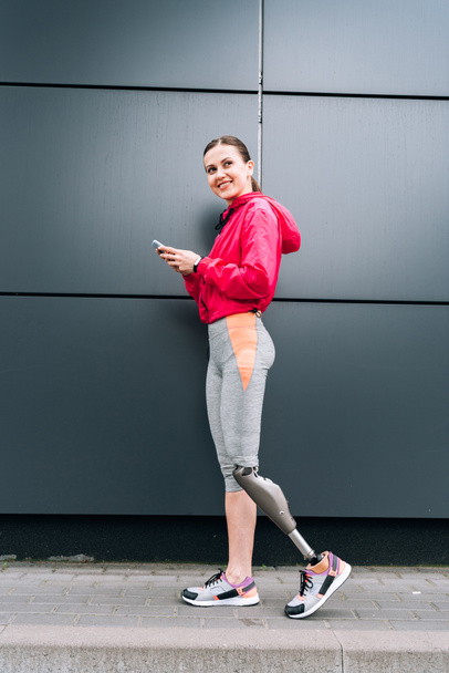 full length view of smiling disabled sportswoman with prosthesis using smartphone on street - Фото, зображення