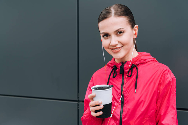 smiling sportswoman holding cup and listening music in earphones on street - Foto, afbeelding