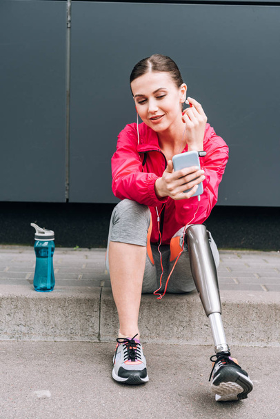 smiling disabled sportswoman listening music in earphones on street - Photo, Image
