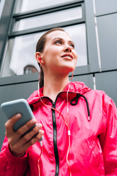 low angle view of smiling sportswoman listening music in earphones and using smartphone on street - Fotoğraf, Görsel