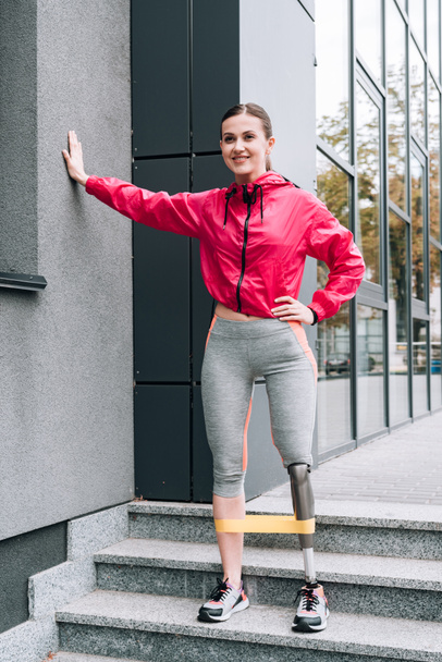 full length view of smiling disabled sportswoman training with resistance band on street - Photo, Image