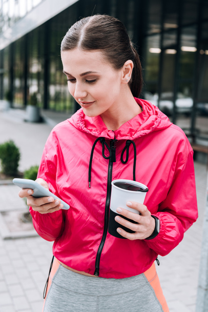 smiling sportswoman holding cup and using smartphone on street - Foto, afbeelding