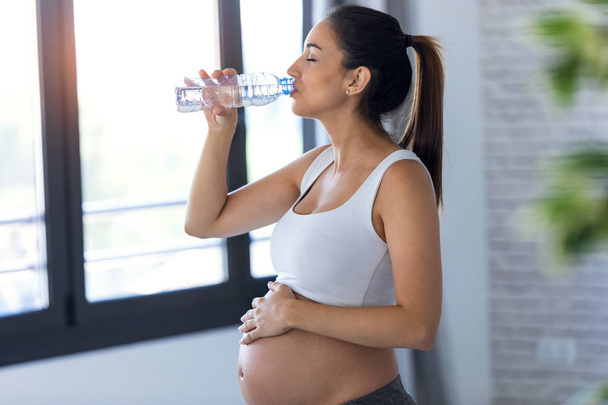 Pretty young pregnant woman drinking water while standing at home. - Фото, зображення
