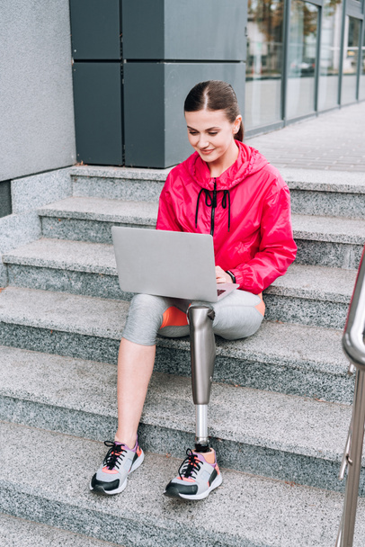 smiling disabled sportswoman using laptop while sitting on stairs on street - Photo, Image