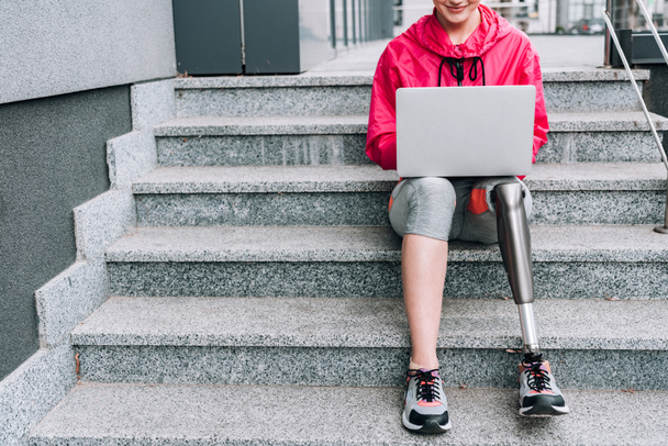 rajattu näkymä hymyilevä vammainen urheilija käyttää kannettavaa tietokonetta istuessaan portaat kadulla
 - Valokuva, kuva