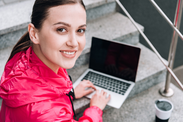 smiling sportswoman using laptop with blank screen on stairs on street - Photo, Image