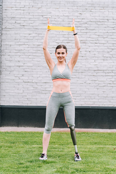 full length view of smiling disabled sportswoman training with resistance band on grass - Photo, Image
