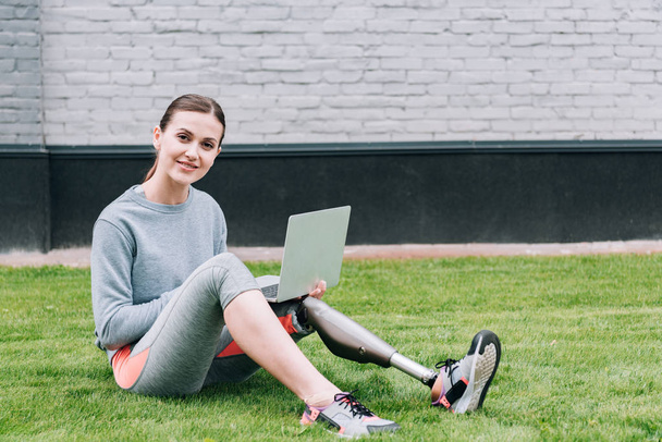 glimlachende gehandicapte sportvrouw met behulp van laptop tijdens het zitten op gras - Foto, afbeelding