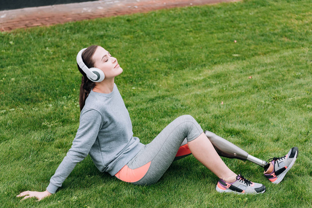 disabled sportswoman sitting on grass and listening music in headphones - Foto, afbeelding