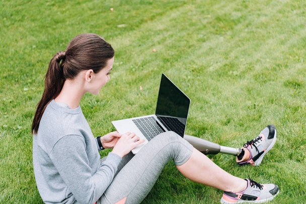 disabled sportswoman using laptop with blank screen while sitting on grass - Photo, image