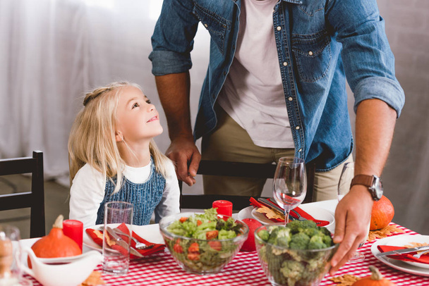 καλλιεργημένη άποψη του πατέρα βάζοντας στο τραπέζι μπολ με μπρόκολο και χαριτωμένο κόρη χαμογελώντας και κοιτάζοντας τον την ημέρα των Ευχαριστιών  - Φωτογραφία, εικόνα