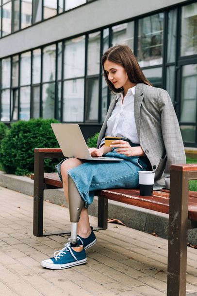 disabled woman with laptop sitting on bench and holding credit card - Foto, Imagem