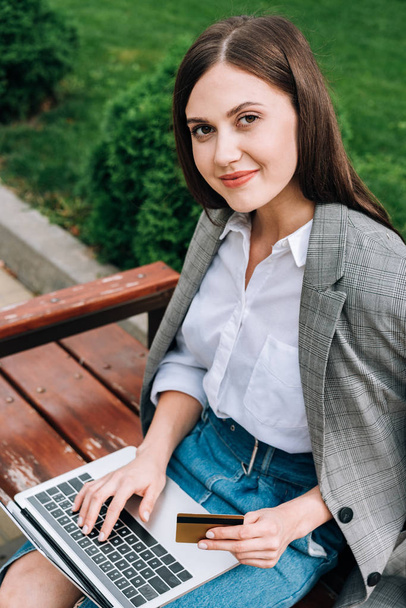smiling young woman with laptop sitting on bench and holding credit card - Фото, изображение