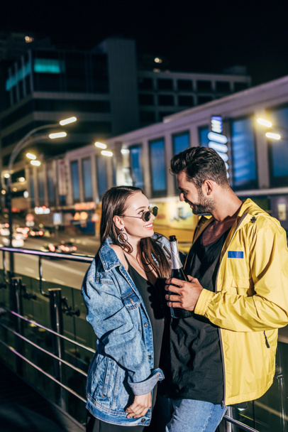 attractive girlfriend and handsome boyfriend with bottle smiling and looking at each other in night city   - Photo, Image