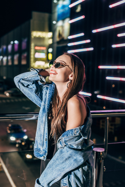 attractive woman in denim jacket and sunglasses smiling and looking away in night city  - Fotoğraf, Görsel