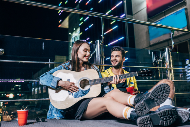 プラスチックカップと夜の街でアコースティックギターを演奏魅力的なガールフレンドとハンサムなボーイフレンド  - 写真・画像