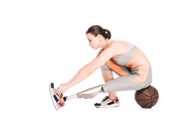 vista lateral de la deportista discapacitada con prótesis sentada sobre pelota de baloncesto y atando cordones aislados sobre blanco
 - Foto, imagen