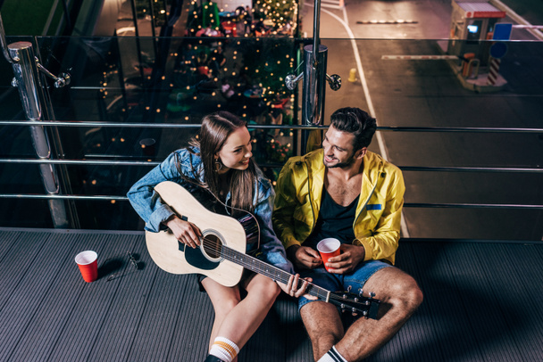 high angle view of handsome boyfriend with plastic cup and attractive girlfriend playing acoustic guitar in night city  - Fotografie, Obrázek